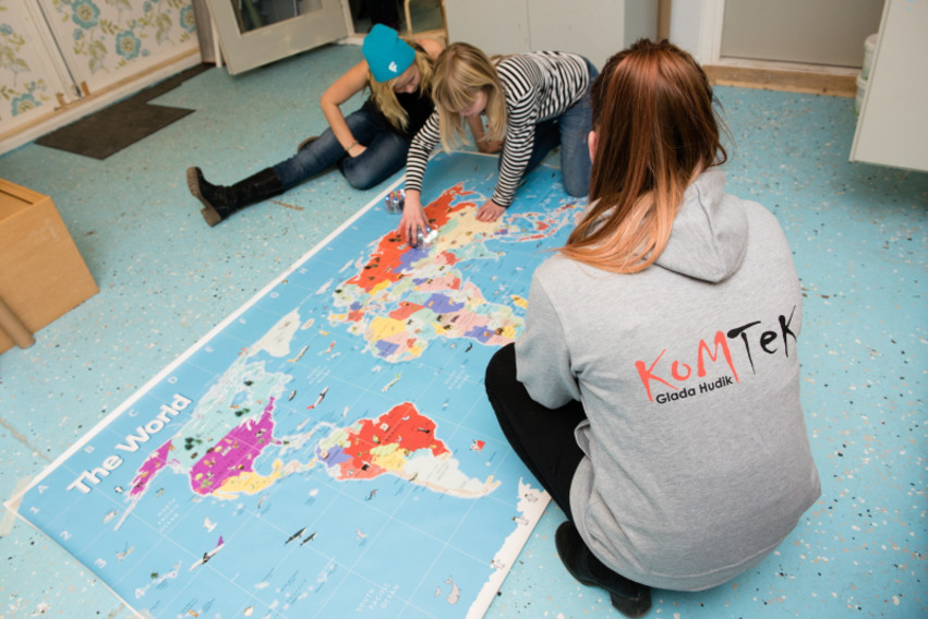 En kvinnlig ledare på KomTek sitter på golvet med två mindre tjejer. Den ena har en blå toppluva på sig. Mellan sig på golvet ligger en jättestor världskarta i papper som en av tjejerna pekar på.