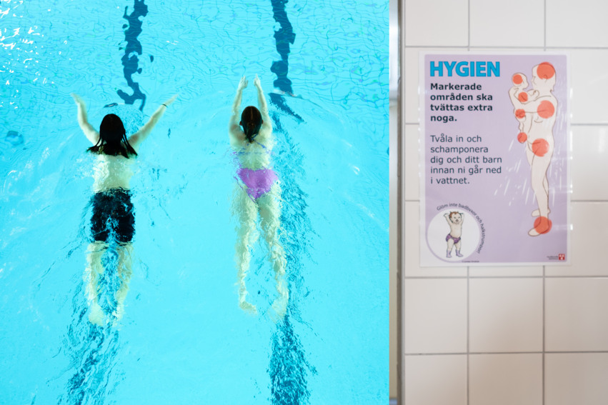 I syns två personer som simmar i en bassäng. Till höger i bild syns en affisch som redogör vilka regler som gäller för hygien på badhusen.