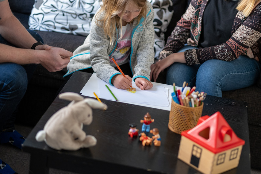 En flicka sitter och målar på ett papper vid ett lågt svart bord. Det sitter två vuxna bredvid henne och på bordet finns det leksaker och en burk med färgpennor.