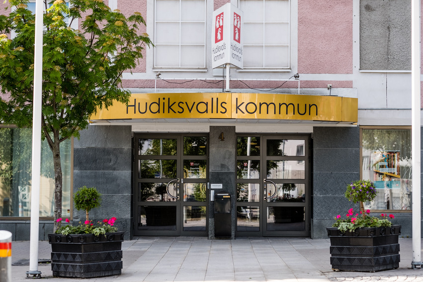 Hudiksvalls kommuns huvudentré. Ovanför entrén sitter en stor guldfärgad skylt där det står Hudiksvalls kommun med stora svarta bokstäver. På varsin sida om entrén står det stora runda blomkrukor med blommor i.