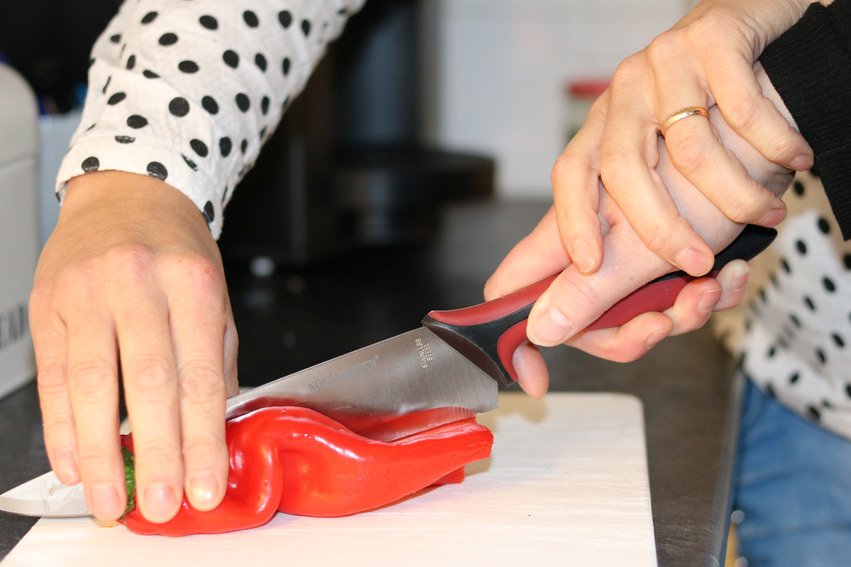 En person hjälper en annan att skära upp en röd paprika på en vit skärbräda. Båda håller i kniven.