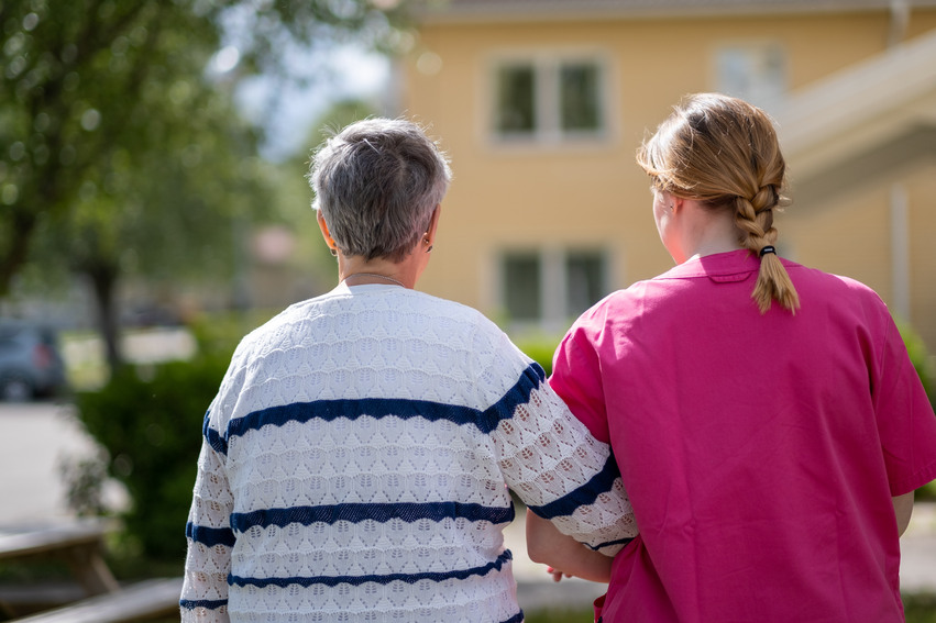 En anställd i sina rosa arbetskläder på äldreboendet tar en promenad med en boende utomhus. De håller varandra under armarna.