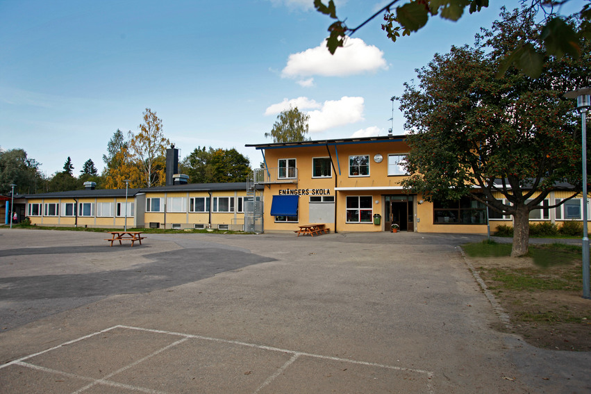 Enångers skola. Byggnaden är avlång och gulmålad. På skolgården står det ett stort träd till höger på en gräsmatta. Träbänkar med bord och en stor asfalterad plan finns utanför skolan.