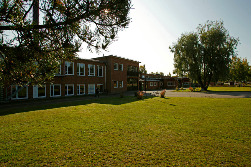 Friggsunds skola. Byggnaderna ligger helt i skugga. På skolgården finns en stor grön gräsmatta som är upplyst av solen. Det finns träbänkar, träd och lekplats.