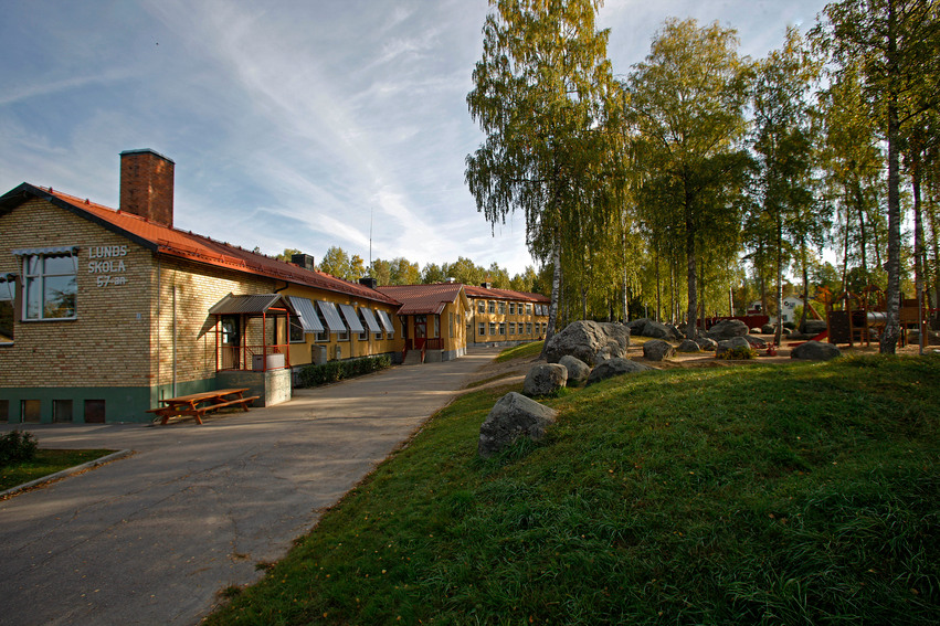 Lunds skola. Skolan består av flera låga byggnader i gult tegel. Till höger finns ett stort skogsområde med lekplats. Stora stenar ligger utspridda.