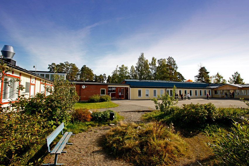 Näsvikens skola. Skolan har flera byggnader i olika färger. På skolgården finns en asfalterad plan och intill den ligger en plats med buskar, planteringar och en bänk att sitta på.