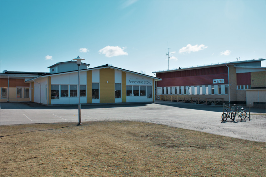 Sandvalla skola. Skolan har flera byggnader i olika färger. Utanför skolan finns en parkering där det står några cyklar. Ett gräsområde med en lyktstolpe finns intill parkeringen.