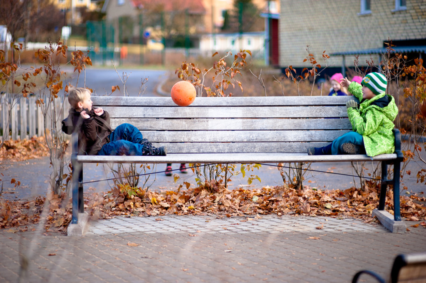 Två barn sitter på en varsin ände av en parkbänk och kastar en orangea boll mellan sig. Under bänken ligger det löv i fina höstfärger.