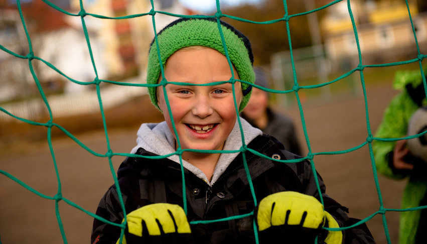 En pojke står inne i ett fotbollsmål och tittar ut mellan de gröna nätmaskorna.