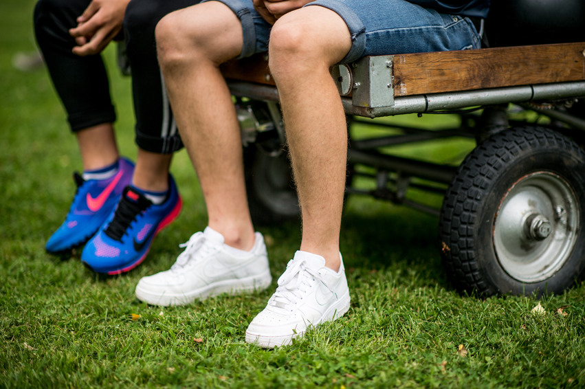 Närbild från midjan och ner på två killar sitter på en vagn med hjul, den står på en gräsmatta. En av dem har vita gympaskor och den andra blåa.