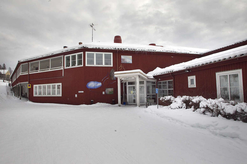 En stor röd byggnad med vita knutar. Entrén är vitmålad och har ett litet tak. Marken och planteringarna utanför är täckta av snö.
