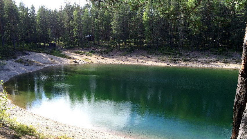 Gröntjärn ligger spegelblank. Vattnet skiftar i blått, grönt och svart och i bakgrunden finns skog med en liten stuga.