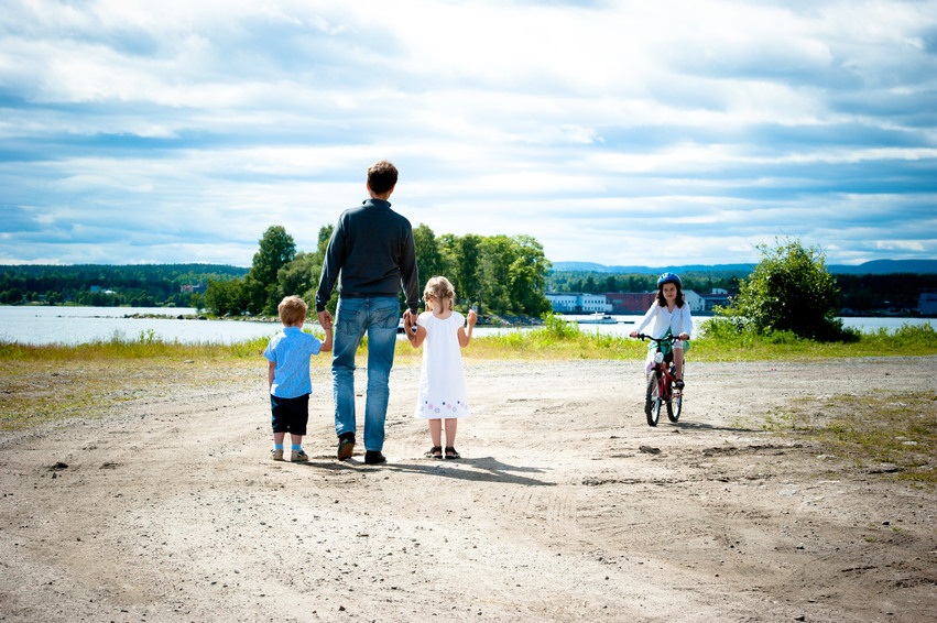 En pappa ute på promenad med sina tre små bar nere vid havet. Ett av barnen cyklar och har en cykelhjälm på huvudet.