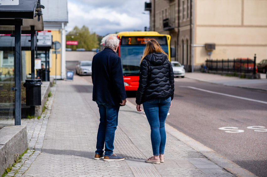 En gammal man och en tjej står tillsammans och väntar på en buss som närmar sig nere på stan.