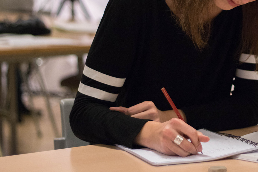 Närbild på en kvinna som sitter vid ett bord och skriver med en blyerspenna. 