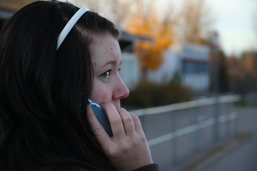 En tjej med långt mörkt hår och ett vitt modem i håret står och pratar i mobiltelefonen samtidigt som hon väntar på tåget.