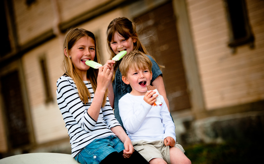 Två små tjejer och en liten kille sitter nere vid Möljen utanför ett av de gulbruna hamnmagasinen och äter glass en fin sommardag.