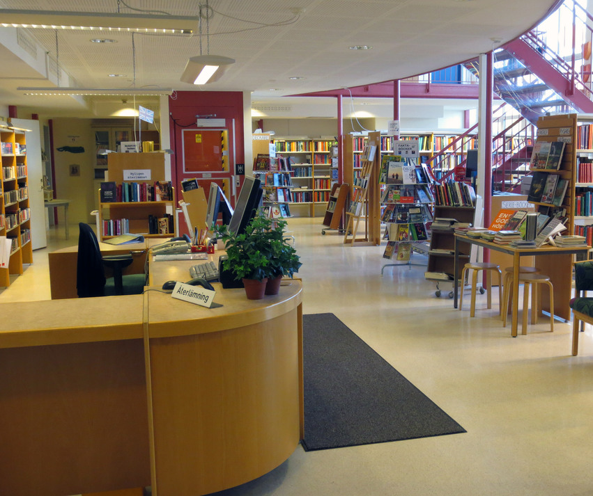 Delsbo bibliotek består av ett stort rum med många bokhyllor som står på rad. En stor gulbrun disk med rundade hörn står till vänster där man kan få hjälp av personalen. En röd trappa leder upp till övervåningen.