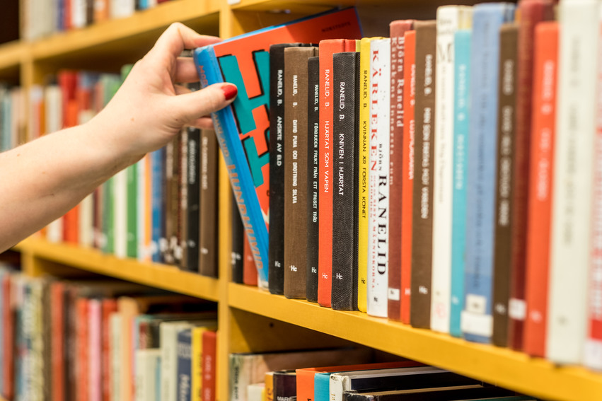 Närbild på en gulmålad bokhylla fylld med böcker i olika färger. En person är precis på väg att ta en bok ur bokhyllan.