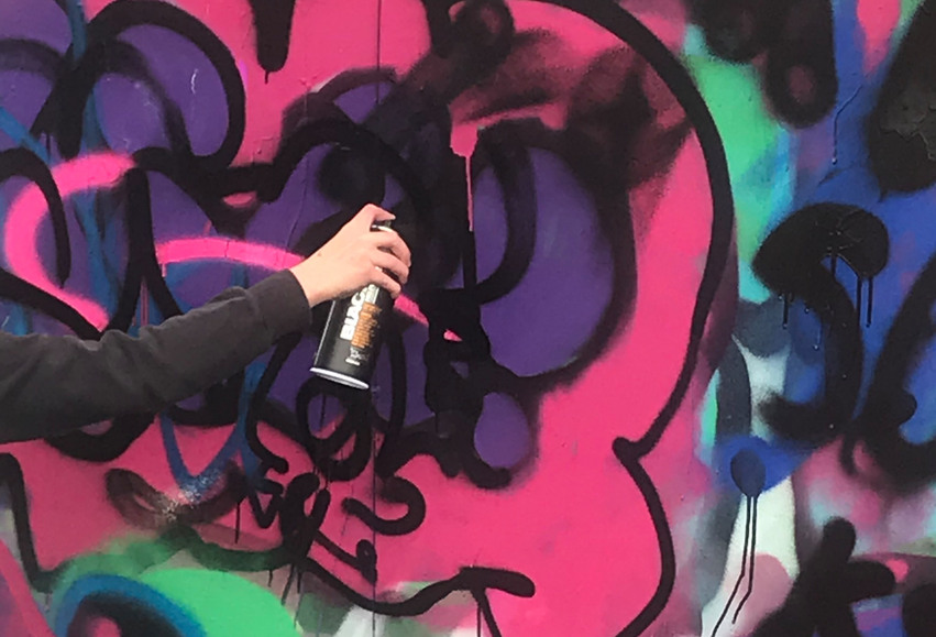 Närbild på en arm och en hand som håller i en sprayburk. Graffitiväggen har ett färgglatt mönster.