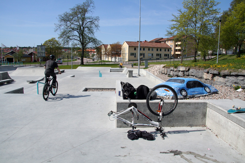 Sportparken en solig dag. En kille cyklar runt bland hindren och en vit cykel ligger upp och ner på betongen utan bakhjul. I bakgrunden ligger gulvita hyreshus med tegeltak.