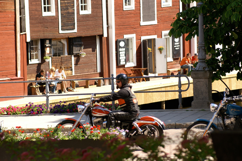 Bakom sjöbodarna finns en lång träbrygga där människor sitter och solar. På vägen bredvid kör två motorcyklister förbi till hälften skymda av blomsterarrangemang.