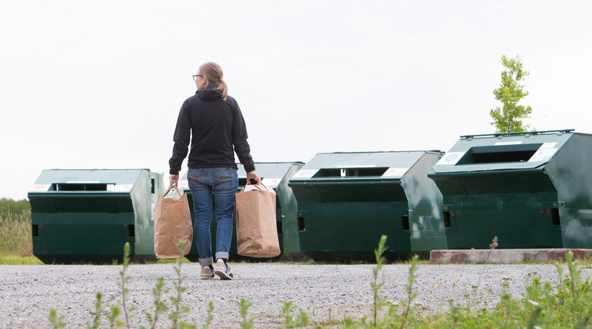 En kvinna går till contianrar på en återvinningsstation för förpackningar och tidningar