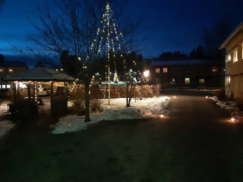 Gårdsplan på Edsbacka vård- och omsorgsboende på kvällen, hus och flaggstång är prydd i julbelysning.
