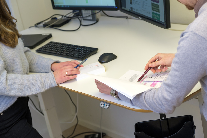 Två personer som står vid ett skrivbord och pekar på ett papper med en varsin penna.