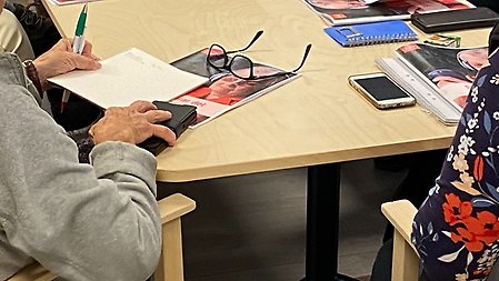 Äldre personer sitter vid ett bord med telefoner, papper och penna framför sig. 