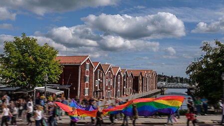 Pridetåg framför sjöbodarna i centrala Hudiksvall. Prideflaggan sveper över folksamlingen.